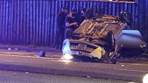 A silver Toyota car, upturned, with a smashed roof and door hanging open. A police officer can be seen stood next to the vehicle.