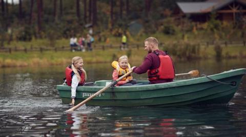 A man in a boat with two children, all wearing lifejackets as they row across a boating pond