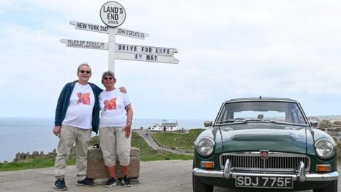 A couple standing under the Land's End finger post and next to a classing car in British Racing Green