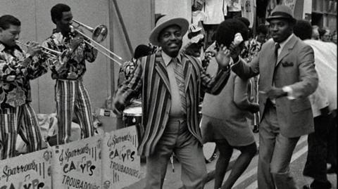 Black and white image of two men in suits dancing in the street to a calypso band.