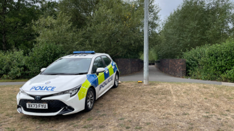 A police car is parked on grass in front of a path over a bridge. There are thick green bushes and trees either side of the path