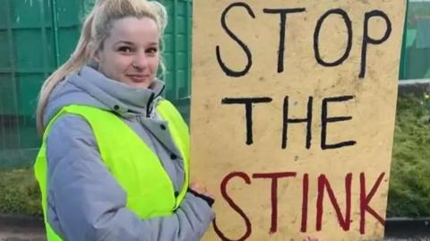 A young woman with long blonde hair, in a grey coat and yellow high vis vest, holds a sign that says 'stop the stink' in front of large green skips at a landfill site.