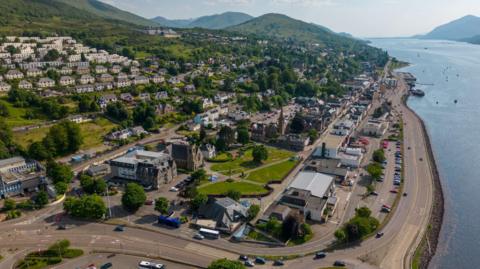 An aerial view of Fort William along a shore of Loch Linnhe on a sunny day. There are industrial and other business premises close to the shore and rows of houses on a hill that rises steeply from the shoreline.