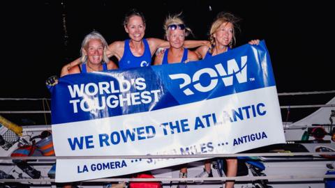 Four women all dressed in blue rowing outfits, standing on the side of a boat and holding a banner that reads "World's Toughest Row". The sky is pitch black in the background. The four women are all smiling. 