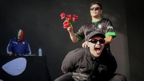 Liam Óg Ó hAnnaidh, aka Mo Chara (C), and Naoise O Caireallain, aka Moglai Bap (R), part of the Northern Ireland hip-hop trio Kneecap, perform on stage at the Rock en Seine music festival, in Saint-Cloud, near Paris, on August 24, 2025.
Mo Chara is seen wearing a black Nike baseball cap, sunglasses and is kneeling forward and yelling animatedly into the camera. Behind him is Moglai Rap, also wearing sunglasses and a Palestine football club shirt, holding a bouquet of six roses. In the background is a third member, wearing an Irish flag balaclava.
