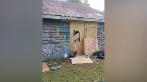 An abandoned building which is boarded up and has graffiti daubed on the walls. It has a tin roof