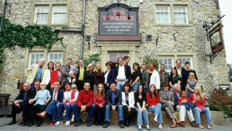 A large group of adults and children pose for a photograph outside a traditional stone-built pub called The Woolpack, with several people seated on hay bales in the front row. The building has a rustic stone façade, sash windows, climbing greenery, and prominent pub signs advertising ales, wine, and food. The group stretches across the entrance and frontage of the pub, creating the appearance of a cast or community gathering in a village setting.
