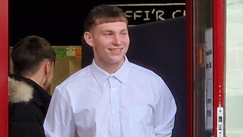 Harvey James with short ginger hair, wearing a white shirt and smiling as he looks off to the side. He is stood in a doorway with a red frame, with another man, wearing a black parka stood behind him with his back to the camera.
