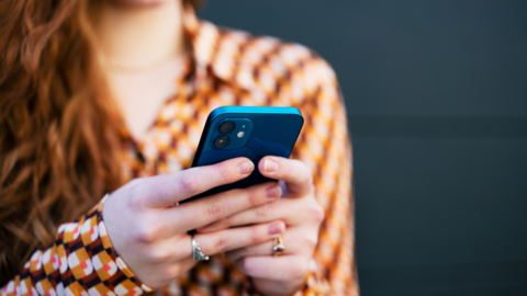 A woman with long red hair and a brown and orange patterned blouse holds a blue smartphone in both hands, as if sending a text message.