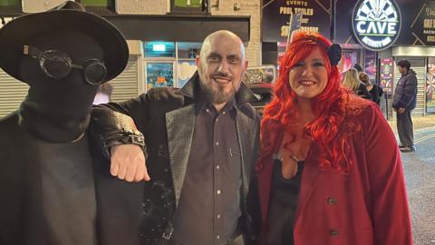 Three people standing in a row wearing fancy dress. The person the left is wearing all black, including their face which is totally covered. They have goggles over the face mask and a hat. The man in the middle is dressed as a vampire and has face paint on. The woman on the right is wearing a red coat and a red hair wig.