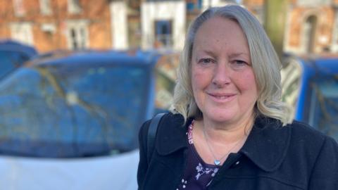 A woman in her 60s who has grey-blond, shoulder-length hair and is wearing a navy blue coat. She is smiling and standing in a car park, with several cars are visible in the background, which is out of focus.