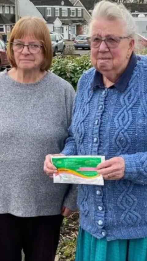 Two elderly women in jumpers outdoors one with red and the other with white hair and one holding a charity shop bag