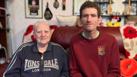 Patricia, with close-cropped silver hair and stud earrings, is wearing a navy Lonsdale jacket, sat next to her son, Colin, with dark brown hair, who is wearing a red jumper. They are sat on a red-coloured sofa with pictures and medals on the wall in the background. 