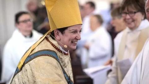 Bishop Anne Dyer dressed in full religious wear, and wearing a small head microphone, smiling while shaking hands with someone. Other people are gathered in background.