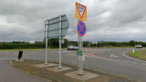 A junction on the A1 with roads joining the main road from both sides. A grey hatchback is driving past on the main carriageway.