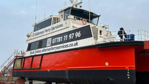 The new ferry is red, black and white with twin-hulls and it has the ferry service phone number and website address displayed on it. The photo shows the boat laid up on hard-standing.