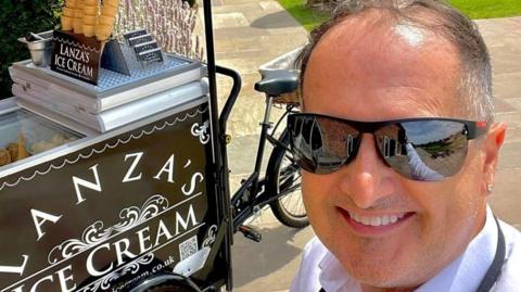 A selfie of a man with a black ice cream cart that is attached to a bicycle. Then man is wearing black sunglasses and smiling. He has short grey hair and a diamond earring in on ear. 