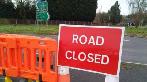 A road closed sign next to a roundabout on a dual carriageway. There are plastic orange barriers. Both have been pulled to the side of the road 