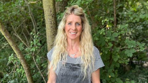 Anna stands in a woodland, smiling at the camera. She has long blonde hair and is wearing a black-and-white striped t-shirt and black denim dungarees.