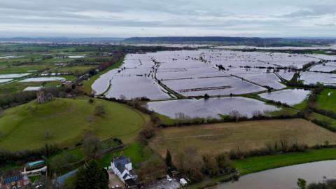 An areal shot of fields covered by water. Glastonbury Tor, a ruin at the top of a hill, is visible to the left of the image. 