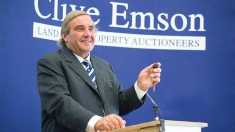 Clive Emson holding a gavel while standing at a lectern. He is wearing a dark thin-striped suit and a striped blue and white tie.