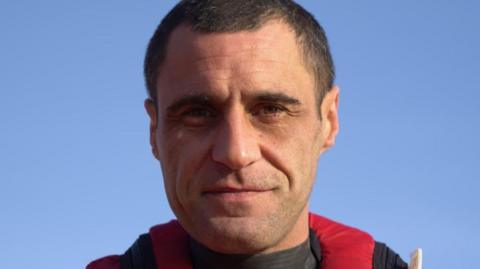 Carl, who has short black hair and very faint stubble, looks at the camera. Behind him is a blue sky