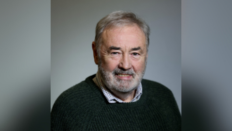John Allen. He has white hair and beard and brown eyes. He is wearing a green jumper with check shirt underneath.