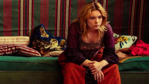 Florence Pugh sits on the end of a bed, looking pensive and anxious.