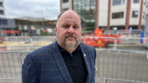 A man with a greying beard and wearing a navy blue jacket looks at the camera with a public area under construction obscured in the background.