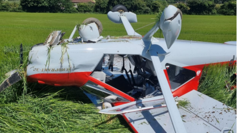 A white and red aircraft which is lying upside in a corn field. There is damage to parts of the plane and grass caught in the wheels and round the propellor.