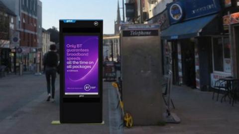 An artist's impression of a large black digital unit with a purple screen on it. It is next to a phone box on a pedestrianised street. 