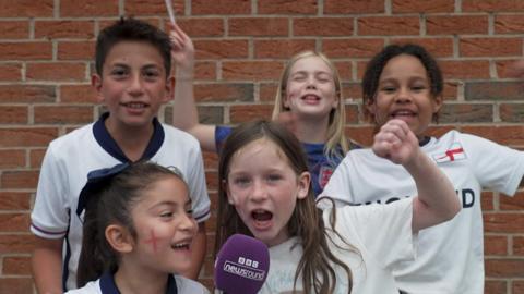 five kids in england kit smiling and cheering in front of a red brick wall with one girl at the front holding a bbc newsround microphone