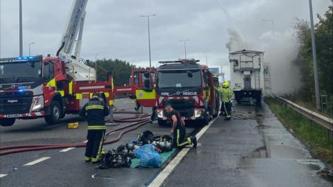 Firefighters battle the flames of a lorry fire on the M5 in Exeter. Two crew members are inspecting breathing apparatus on the road while others work on the fire.