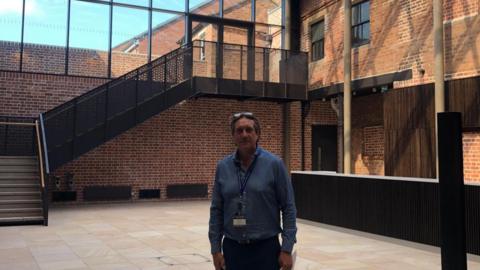 Matt Smith stood in the new bar area with exposed brick on the walls and a large black staircase behind him. He is wearing a blue shirt with a blue lanyard and sunglasses on his head. 
