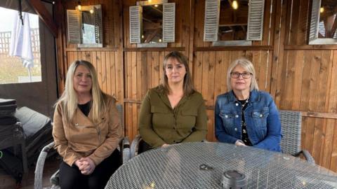 Three women sat in a row, first has blonde hair and is wearing a light brown jacket, the second has darker hair with a fringe and is wearing a green shirt and the third has short grey hair that comes to her shoulders and is wearing glasses and a denim jacket. They are sat in an outdoor seating area with wooden panels behind them.