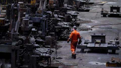 A wide view inside an industrial steelworks, showing long rows of large, dark metal machinery and workstations. A worker in bright orange protective clothing walks along the factory floor between the machines, with cables, metal parts, and heavy equipment scattered across the ground under dim industrial lighting.