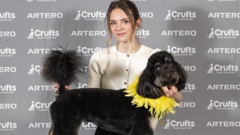 A young woman wearing a white blouse standing behind a black cockapoo with white patches on its legs and a yellow decoration around its neck. Both are looking at the camera.