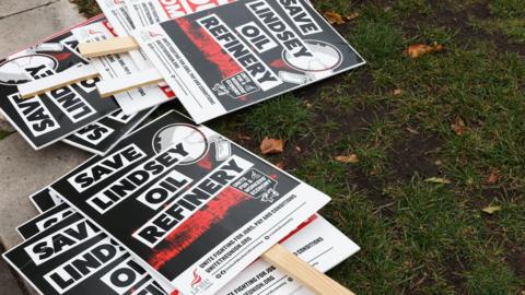 Black and white placards on wooden sticks on the ground outside parliament in London. The placard include a slogan reading "Save Lindsey Oil Refinery" and the logo of the Unite union. They have been laid on paving stones and green grass.