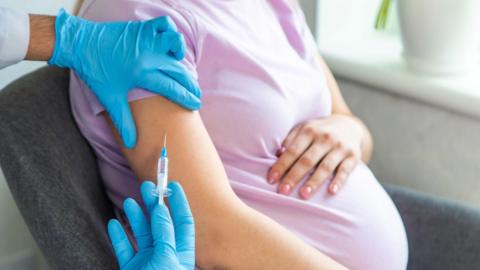 The image is a close up of a pregnant woman who is wearing a pink t-shirt. She is sitting on a grey chair and has her left hand over her pregnant belly. A medical professional, who is wearing blue latex gloves, is holding her right arm with their left hand and is holding a needle near the woman's arm.