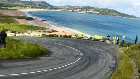 A corner of the track at the Donegal International Rally, overlooking a beach, with a handful of spectators standing behind the barrier 