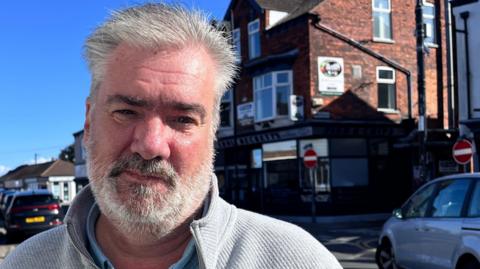 Ian Stead stands in front of a road and parked cars in Market Place, Cleethorpes. He has grey hair and a grey beard and wears a light-grey cardigan. Shops can be seen in the background.