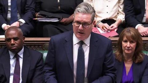 Sir Keir Starmer in a dark blue suit, white shirt and dark blue tie has greying slick backed hair and black spectacles. He is flanked by David Lammy on the left who has greying hair and a similar outfit. Lammy is Afro-Caribbean. Rachel Reeves is on his right - she has long brunette hair and wears a dark blue suit and bright blue blouse.