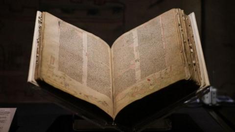 A medieval book open on a stand on a table, which a black background. The book has yellowing pages covered with five columns of densely written text and drawings around the edges.