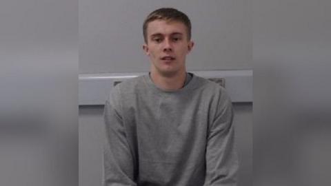 A young man with short brown hair, pictured in a grey sweatshirt against a grey wall