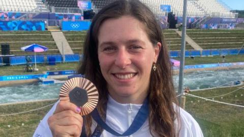 Kimberley Woods holding her bronze medal and smiling at the camera. Behind her is a kayaking course and a block of seats, which are all empty