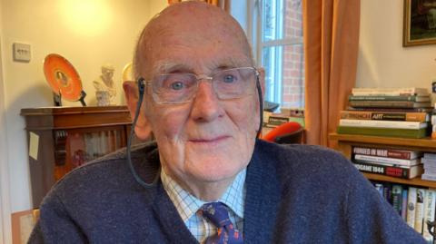 Denis Kenyon wearing glasses, a tie, shirt and blue jumper. He is looking at the camera with books in the background.
