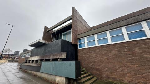 A general view of the front of the former crown court in Ipswich. It is a red brick building and its front entrance has been boarded up with large black boards. The windows on the building have blinds pulled across them. 