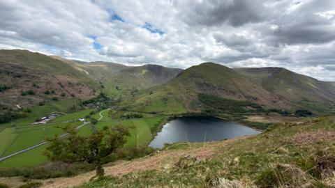Thick clouds over a view of the Cumbrian fells, with a flat, calm lake visible in the foreground.