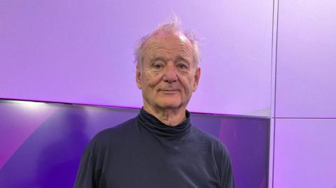Bill Murray pictured in the BBC Radio WM studio wearing a navy blue turtleneck jumper. He has thin, white hair. He is standing in front of a TV screen which has purple BBC Radio WM branding on it. 