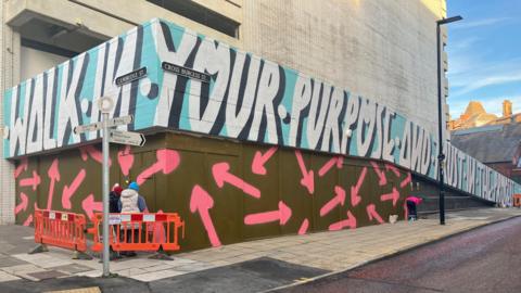 The lettering is on the barrier running down the side of a car park ramp. It it white with black shadowing which makes it appear 3D. The background is light blue. Below it, on a green area of wall or barrier, lots of pink arrows have been painted.
